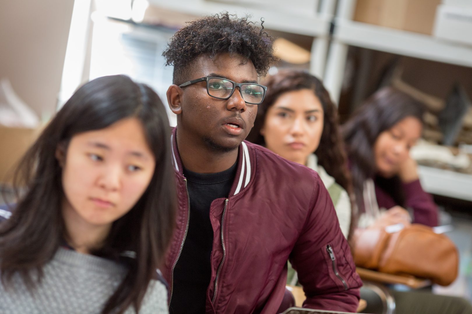 Group of diverse students seated in a classroom or library; a young man with curly hair and glasses in a maroon jacket is in the foreground, looking attentive.